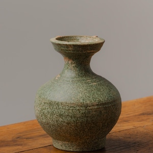 May include: A green ceramic vase with a rounded body and a narrow neck. The vase has a speckled texture and a slightly flared rim. The vase is displayed on a wooden surface.