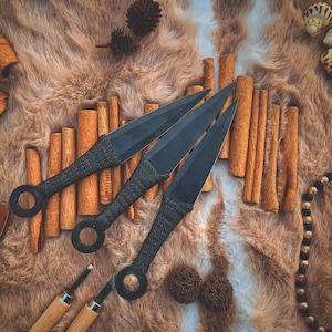 May include: Three black throwing knives with woven handles and ringed pommels are displayed. The knives are arranged on a textured, brown surface with cinnamon sticks. The blades are sharp and pointed, suggesting a focus on craftsmanship and utility.