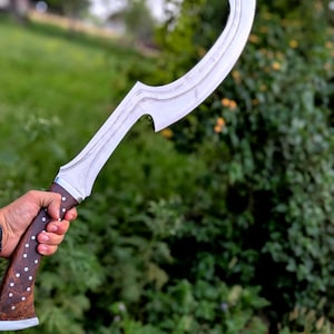 Op de afbeelding: Een gebogen, zilverkleurig mes met een houten handvat. Het handvat is donkerbruin met witte stippen. Het mes heeft een kenmerkende vorm, met een gebogen rand en een punt. De achtergrond is wazig, met groen gebladerte.