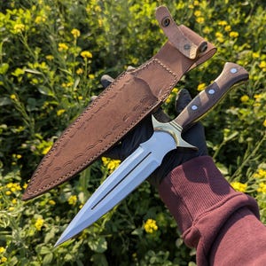 May include: A double-edged dagger with a polished steel blade and a wooden handle with brass accents. The dagger is displayed with its brown leather sheath, which has decorative stitching. The background is a field of yellow flowers.