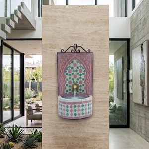 May include: A decorative wall fountain with a mosaic design in a modern home. The fountain features a pink and green geometric pattern, with water flowing from a golden spout. The surrounding walls are a light beige color.