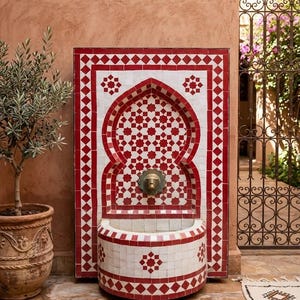 Op de afbeelding: Decoratieve wandfontein met een rood en wit tegelontwerp. De fontein heeft een gewelfde bovenkant en een bekken, met een messing kraan. De tegels zijn gerangschikt in geometrische patronen.