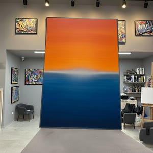 Puede incluir: Gran pintura abstracta con un degradado de tonos naranja y azul, dividida por una suave línea blanca. La obra está enmarcada en negro y se apoya contra una pared en un estudio. Otras obras enmarcadas son visibles en el fondo.