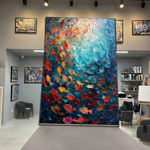 Könnte beinhalten: Großes abstraktes Gemälde, das einen Fischschwarm in Rot-, Orange- und Gelbtönen vor einem strukturierten blauen Hintergrund darstellt. Das Kunstwerk wird in einer Galerie ausgestellt, mit anderen gerahmten Kunstwerken.