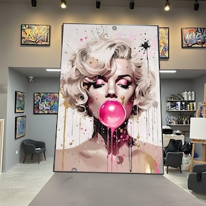 May include: Large framed artwork featuring a pop art style portrait of Marilyn Monroe blowing a pink bubble. The artwork has a white background with pink and gold paint splatters. The subject has closed eyes and pink eyeshadow.