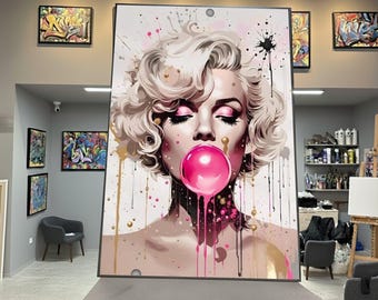 May include: Large framed artwork featuring a pop art style portrait of Marilyn Monroe blowing a pink bubble. The artwork has a white background with pink and gold paint splatters. The subject has closed eyes and pink eyeshadow.