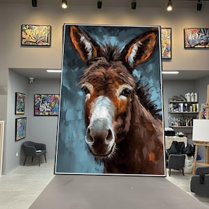 May include: Large framed oil painting of a donkey's face. The artwork features a close-up of the animal's head, with brown and white fur and large ears. The background is a textured blue. Other framed art pieces are visible in the background.