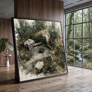 May include: A large square framed wall art piece depicting a high-angle view of a secluded outdoor fire pit area surrounded by large grey boulders, green trees, and wild grasses, set within a room with wood paneling and a large window.