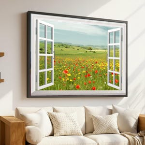 Op de afbeelding: Een ingelijst kunstwerk dat een open raam afbeeldt met uitzicht op een veld vol rode en gele wilde bloemen. Het zwarte frame omlijst witte ramen, die een groen landschap onder een blauwe lucht tonen.
