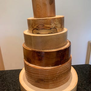 May include: A collection of wooden discs, stacked to form a tower. The wood varies in colour from light to dark brown, highlighting the natural grain. The arrangement resembles a rustic sculpture or cake, with the largest disc at the base.