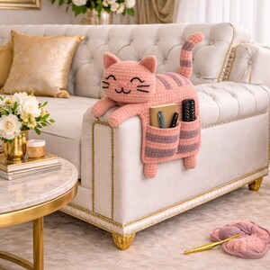 May include: A pink crocheted cat-shaped couch arm tidy with a smiling face and striped tail. The organiser has pockets for storing items like pens and remote controls. The cat is resting on a white couch with gold trim.
