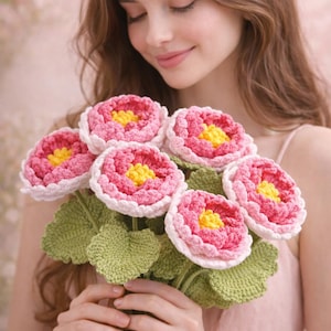 May include: A bouquet of handmade crochet flowers. The flowers have pink petals with white edges and yellow centers. The green leaves and stems complete the floral arrangement. The bouquet is held in front of a soft pink background.