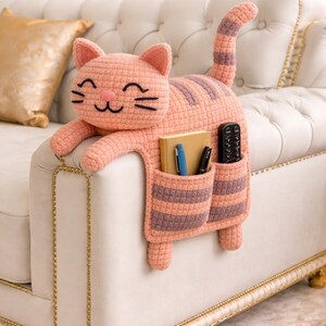 May include: A peach-coloured, crocheted cat-shaped couch organiser with striped pockets. The cat has a smiling face and holds a book, pens, and a remote. The organiser is designed to drape over the arm of a couch.