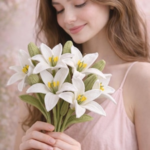 May include: A bouquet of white crochet lilies with yellow centers and green stems and leaves. The handmade flowers are held in front of a person wearing a pink top.