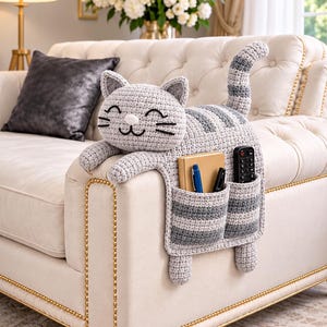 以下が含まれることがあります： グレーと白のストライプ柄のクロッシェ編みの猫型ソファアームレスト用オーガナイザー。猫は刺繍された目とひげを持つ笑顔の顔、長い尻尾、ノート、ペン2本、黒いリモコンが入った2つのフロントポケットを備えています。