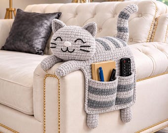 May include: A gray and white striped crochet cat organizer draped over a sofa arm. The cat features a smiling face with embroidered eyes and whiskers, a long tail, and two front pockets containing a notebook, two pens, and a black remote control.