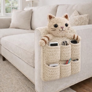May include: A beige crochet cat-shaped organizer with multiple pockets, designed to hang over the arm of a sofa. The cat has brown accents and holds pockets containing items like phones and glasses. The organizer is made of a textured, soft material.