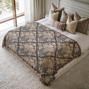 May include: A rectangular woven throw blanket featuring a dark charcoal grey background with a repeating cream-coloured floral damask pattern. The blanket has a fringed edge with multi-coloured threads and is draped over a white bed with several pillows.