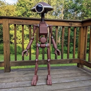 Op de afbeelding: Een bruine, humanoïde robotfiguur met een groot, cirkelvormig oog met een donkere lens in een koepelvormig hoofd. De robot heeft een slank, gearticuleerd frame met zichtbare gewrichten bij de schouders, ellebogen, heupen en knieën, eindigend in klauwvormige handen.