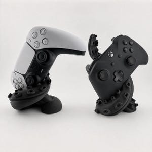 Puede incluir: Un mando de videojuego blanco y negro a la izquierda y un mando de videojuego negro a la derecha, cada uno sostenido por un soporte de tentáculo de pulpo negro con ventosas visibles, diseñado para exhibir hardware de juego en una superficie.