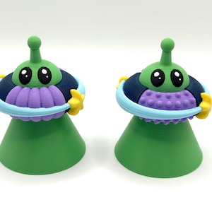 May include: Two green, purple, and blue alien figurines. Each has a green head with large black eyes and a small antenna. The alien is wearing a blue and purple ring with yellow star-shaped hands. The base is green.