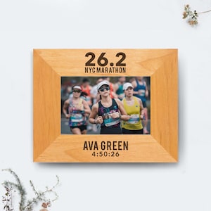 May include: A wooden picture frame with a photo of runners in a marathon. The text "26.2 NYC MARATHON" is at the top, and "AVA GREEN 4:50:26" is at the bottom. The frame is a light brown color.