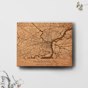 May include: A wooden map of Philadelphia, PA, with intricate street and river details. The city's name and coordinates are engraved at the bottom. The wood has a natural, light brown color.