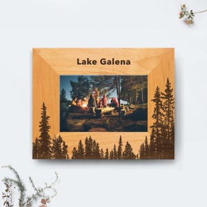 May include: A wooden picture frame with the text "Lake Galena" at the top. The frame has a photo of people around a campfire. The frame has a forest silhouette design on the bottom and sides.
