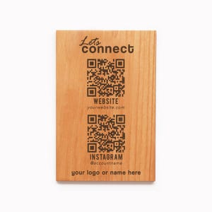 Peut inclure: Panneau rectangulaire en bois clair avec le texte "Lets connect". Deux codes QR sont affichés, un pour un site web et un pour Instagram. Le nom d'utilisateur Instagram est affiché en dessous. Le bas du panneau indique "your logo or name here".