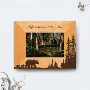 May include: A rectangular light-colored wooden picture frame featuring a dark brown laser-engraved silhouette of a large bear with two cubs and pine trees. The top of the frame displays the text Life is better at the cabin in dark brown.