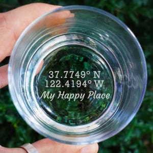 May include: Clear glass tumbler with the text "37.7749° N 122.4194° W My Happy Place" etched on the bottom. The glass is held up, with a blurred green background visible through the glass.
