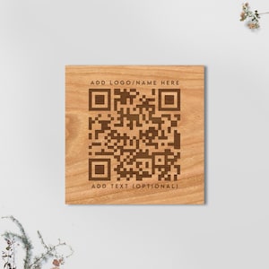May include: Square wooden plaque with a QR code and the text "ADD LOGO/NAME HERE" and "ADD TEXT (OPTIONAL)". The QR code and text are in a dark brown color, set against a light brown wood grain background.