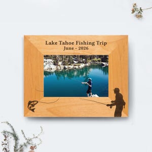 Könnte beinhalten: Holzbilderrahmen mit einem Foto von einer Person, die an einem See angelt. Der Rahmen ist mit einem Fisch und einer Fischer-Silhouette graviert. Der Text "Lake Tahoe Fishing Trip June - 2026" ist oben aufgedruckt.