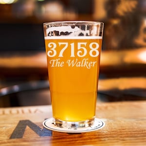 Könnte beinhalten: Ein Pintglas gefüllt mit goldenem Bier, bedeckt mit Schaum. Auf dem Glas steht in weißer Schrift "37158 The Walker". Das Glas steht auf einem Untersetzer auf einem Holztisch.
