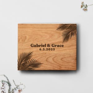 May include: A wooden square with engraved text and palm frond designs. The text reads "Gabriel & Grace" above "6.5.2025". The wood has a warm, natural tone, and the palm fronds are dark brown.
