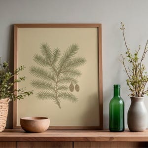 May include: A framed print featuring a detailed illustration of a pine branch with pinecones in shades of green and brown. The artwork is displayed on a wooden surface, alongside a green bottle, a vase, a wooden bowl, and a woven basket.