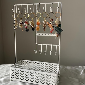 May include: A white metal jewelry organizer with multiple hooks and a basket base. Various keychains with colorful charms hang from the top. The organizer is designed to display and store jewelry and accessories.