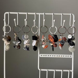 May include: A collection of handmade keychains hanging on a white metal display rack. Each keychain features a unique assortment of decorative elements, including buttons, charms, and beads in various colors and shapes. The keychains are attached to silver key rings.
