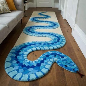 May include: A blue snake-shaped rug with a mosaic pattern in shades of blue. The rug's body winds across a cream-colored runner, with a detailed snake head featuring a red tongue. The rug is placed on a wooden floor.