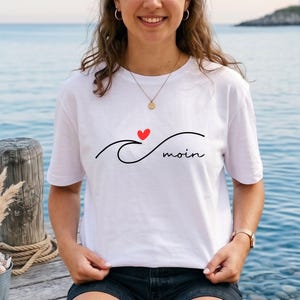 Könnte beinhalten: Weißes T-Shirt mit einem schwarzen Wellen-Design, einem roten Herz und dem Wort "moin" in schwarzer Schrift. Das kurzärmelige Shirt scheint aus Baumwolle zu sein.