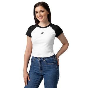 Może przedstawiać: Biały top z czarnymi rękawami raglan i małym czarnym logo na piersi. Modelka ma na sobie niebieskie dżinsy. Koszulka jest w stylu casual.