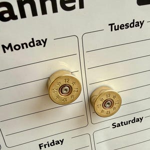May include: Two brass bullet casing magnets on a white planner. The magnets are cylindrical with the number "12" stamped on the rim. The planner has the days of the week printed on it, including "Monday", "Tuesday", "Friday", and "Saturday".