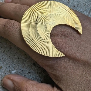 May include: A large, gold-colored ring in a crescent moon shape. The ring has a textured, sunburst design radiating from the center. The jewelry is worn on a hand, showcasing the ring's size and design.