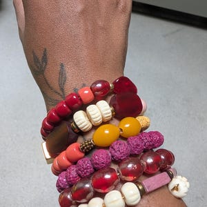 Può includere: Una pila di braccialetti di perline con una varietà di forme, dimensioni e consistenze. Le perline includono vetro liscio rosso, arancione e giallo, pietre laviche magenta porose, forme floreali bianco sporco e una pietra rettangolare rosa.