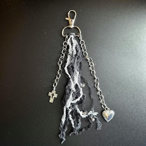 May include: A silver-toned metal keyring featuring a lobster clasp, two dangling chains, and a central cluster of black and white lace ribbons. One chain ends with a small cross charm, while the other chain features a small heart-shaped locket.