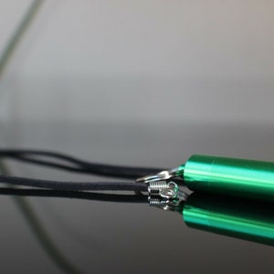 May include: A green metal pill case with a black cord attached. The case is cylindrical and has a rounded end. The cord is looped around the case and has a silver clasp.