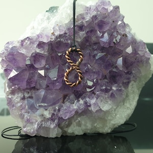 May include: A copper infinity symbol pendant on a black cord, resting on a cluster of amethyst crystals.