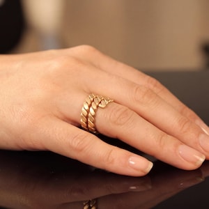 May include: A gold-colored ring set on a hand. The ring has a braided rope design. The ring is worn on the ring finger. The hand is resting on a dark surface.