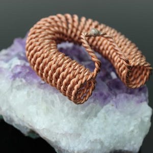 May include: A copper wire coil, shaped in a U-shape, rests on a piece of purple and white stone. The coil is made of tightly wound copper wire, and the stone has a crystalline structure.