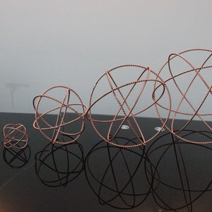 Puede incluir: Un conjunto de cinco adornos esféricos de alambre de cobre de diferentes tamaños. Las esferas están hechas de alambre fino y están dispuestas en línea sobre una superficie negra.